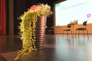 Das Schmuckbild zeigt einen geschmückten Blumenkübel vor den noch unbesetzten Tischen für den Eröffnungstalk und im Hintergrund eine Leinwand mit dem Schrift #Viersenbluehtauf (Foto: Stadt Viersen)