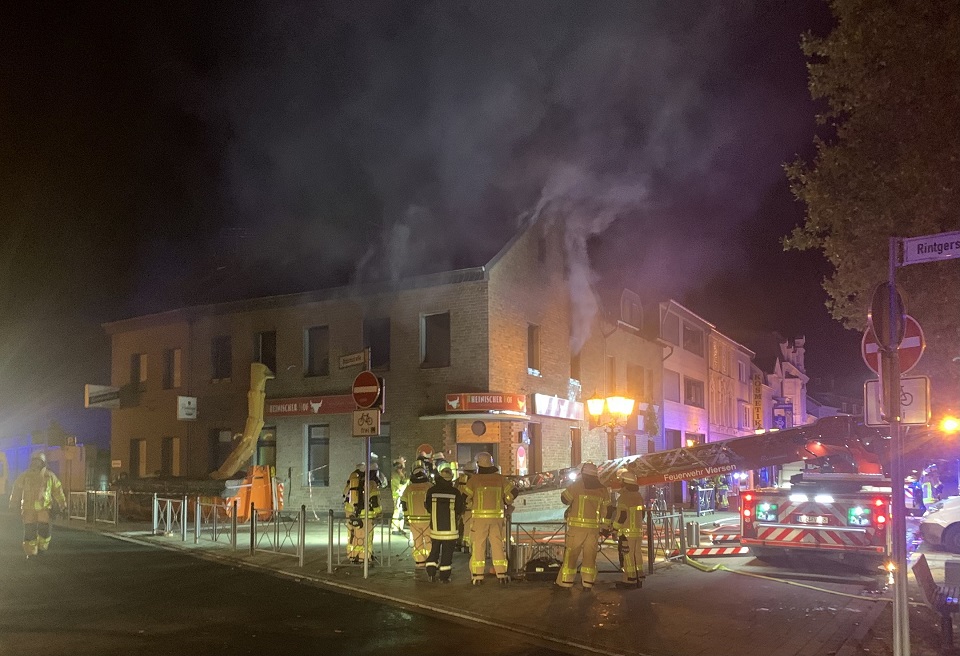 Feuerwehrkräfte im Einsatzanzug vor einem Wohnhaus, Rauch dringt durch Fenster, städtische Beleichtung und Scheinwerfer erhellen die Nacht 